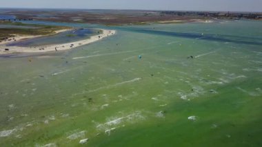 Aerial of many kiteboarders with colorful kites flying over the blue sea lagoon ride on kiteboards. Kitesurfers surf on waves with parachutes on a windy day. Drone video.
