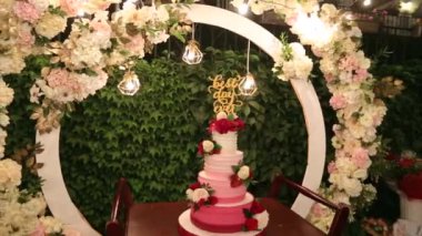 Wedding cake with best day ever sign on green garden decorated for wedding party at night. White, marsala creative decor with circle wedding arch decorated with roses and flowers, fairy lights.