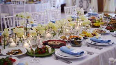 Wedding rustic style decor with dishes, drinks and roses in white and blue colours on table setting at restaurant. Banquet hall interior floristic details, flowers in glass vases filled with water.