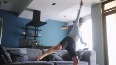 Young futuristic blonde girl wearing virtual reality headset and holding controller playing tennis video game at home. Woman playing VR tennis competition in metaverse in the middle of living room.