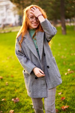 Redhead smiling pretty woman walking in park on sunny autumn day. Elegant female poses in stylish authentic outfit has fun laughing on green lawn with vivid foliage, leaves. Girl enjoys a fall season.