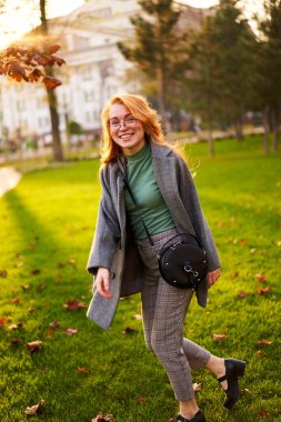 Redhead smiling pretty woman walking in park on sunny autumn day. Elegant female poses in stylish authentic outfit has fun laughing on green lawn with vivid foliage, leaves. Girl enjoys a fall season.