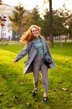 Redhead smiling pretty woman walking in park on sunny autumn day. Elegant female poses in stylish authentic outfit has fun laughing on green lawn with vivid foliage, leaves. Girl enjoys a fall season.