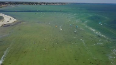 Aerial of many kiteboarders with colorful kites flying over the blue sea lagoon ride on kiteboards. Kitesurfers surf on waves with parachutes on a windy day. Drone video.