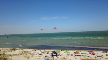 Aerial of many kiteboarders with colorful kites flying over the blue sea lagoon ride on kiteboards. Kitesurfers surf on waves with parachutes on a windy day. Drone video.