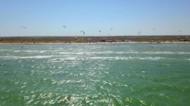 Aerial of many kiteboarders with colorful kites flying over the blue sea lagoon ride on kiteboards. Kitesurfers surf on waves with parachutes on a windy day. Drone video.