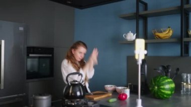 Food blogger woman cooking fresh vegan salad of vegetables in kitchen studio and filming funny dancing for social media content on a smartphone for a video channel. Female influencer streaming.