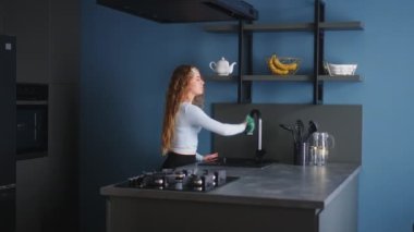 Pretty young caucasian woman wearing headband wipes surfaces at home in a modern stylish kitchen, keeping house neat. Housewife cleaning kitchen surface with rag. Young female doing household chores