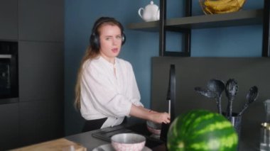Authentic happy caucasian woman in a headphones washing the dishes and dancing in a modern kitchen. Young housewife listening music, having fun while doing the washing-up.