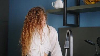 Authentic happy caucasian woman washing the dishes, singing and dancing in modern kitchen. Young housewife having fun while doing washing-up. Girl waving her hair while doing the dishes