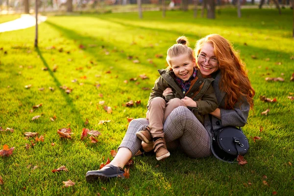 Smiling redhead woman and little boy sitting on a grass lawn in city park on warm autumn day. Young mom hugs her son, they have fun and look at camera on sunny fall day. Foliage on green lawn.