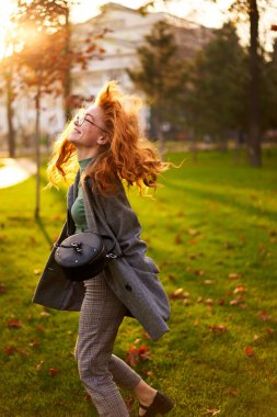 Redhead smiling pretty woman walking in park on sunny autumn day. Elegant female poses in stylish authentic outfit has fun laughing on green lawn with vivid foliage, leaves. Girl enjoys a fall season.