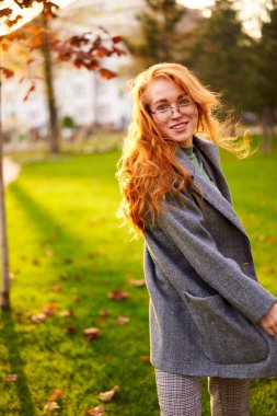 Redhead smiling pretty woman walking in park on sunny autumn day. Elegant female poses in stylish authentic outfit has fun laughing on green lawn with vivid foliage, leaves. Girl enjoys a fall season.