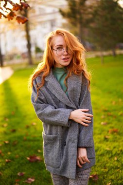 Redhead smiling pretty woman walking in park on sunny autumn day. Elegant female poses in stylish authentic outfit has fun laughing on green lawn with vivid foliage, leaves. Girl enjoys a fall season.