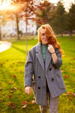 Redhead smiling pretty woman walking in park on sunny autumn day. Elegant female poses in stylish authentic outfit has fun laughing on green lawn with vivid foliage, leaves. Girl enjoys a fall season.