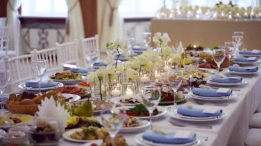 Wedding rustic style decor with dishes, drinks and roses in white and blue colours on table setting at restaurant. Banquet hall interior floristic details, flowers in glass vases filled with water.