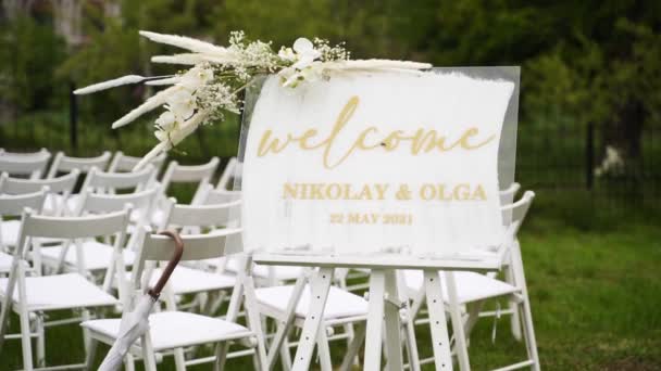 白いバラ 天然の花で飾られた屋外結婚式のための結婚式歓迎サイン装飾 緑の芝生の上でお客様のための白い椅子 夏の田舎の結婚式の花屋のデザイン — ストック動画