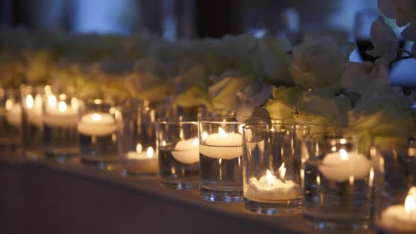 ガラスの花瓶に浮かんでいるキャンドルは水で満たされたフラスコ 宴会場の内部の花の詳細 パーティーライトだ レストランでの新婚旅行のダイニングテーブルの設定で白いバラと結婚式の装飾 — ストック動画