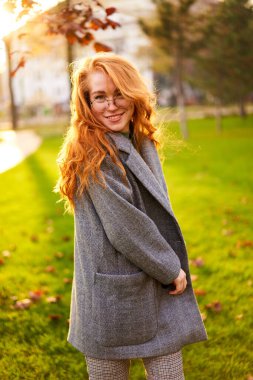 Redhead smiling pretty woman walking in park on sunny autumn day. Elegant female poses in stylish authentic outfit has fun laughing on green lawn with vivid foliage, leaves. Girl enjoys a fall season.