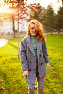 Redhead smiling pretty woman walking in park on sunny autumn day. Elegant female poses in stylish authentic outfit has fun laughing on green lawn with vivid foliage, leaves. Girl enjoys a fall season.