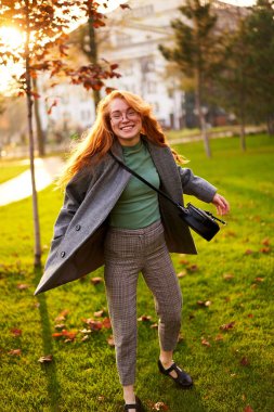 Redhead smiling pretty woman walking in park on sunny autumn day. Elegant female poses in stylish authentic outfit has fun laughing on green lawn with vivid foliage, leaves. Girl enjoys a fall season.