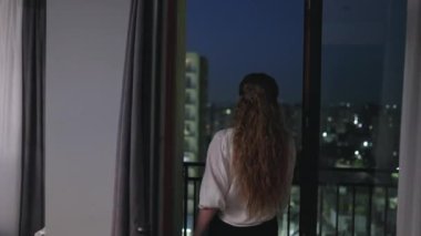 Young woman walking out from apartment enjoying night city view. Girl rests on balcony balustrade. Pretty lady looking at lights. Female going through glass door. Observing street lights at terrace