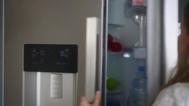 Woman putting groceries in a new modern fridge. Young adult female unpacking shopping bag loading modern hi-tech refrigerator. Girl putting fruit and vegetables into fridge closeup shot
