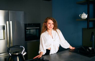 Portrait of a blonde caucasian woman standing and smiling in her kitchen leaning on the table. Young cheerful adult female standing at home in modern interior with hands on cooking surface