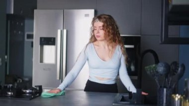 Pretty young caucasian woman wearing headband wipes surfaces at home in a modern stylish kitchen, keeping house neat. Housewife cleaning kitchen surface with rag. Young female doing household chores