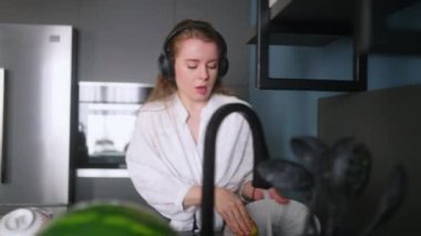 Authentic happy caucasian woman in a headphones washing the dishes and dancing in a modern kitchen. Young housewife listening music, having fun while doing the washing-up.