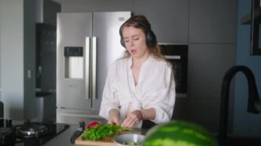 Woman in headphones making a salad of vegetables and has fun dancing at modern kitchen island. Young female cuts the greens and cooks salad. Girl cuts cucumbers and tomatoes on a chopping board.