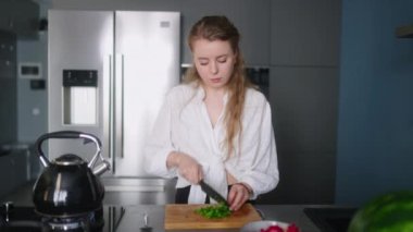 Caucasian woman making a salad of vegetables and tasting a meal on a stove on modern kitchen island. Young female cuts greens and cooks a dish. Girl cuts cucumbers and tomatoes on chopping board.
