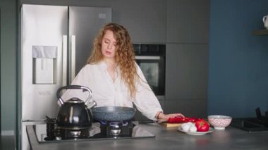 Young curly woman cutting tomatoes on chopping board to make an omelette on a pan. Young female cooking her meal. Girl preparing her breakfast on gas stove of a modern minimalistic kitchen island.
