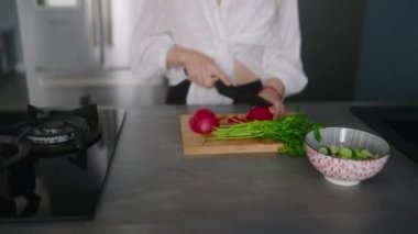 Woman in headphones making a salad of vegetables and has fun dancing at modern kitchen island. Young female cuts the greens and cooks salad. Girl cuts cucumbers and tomatoes on a chopping board.