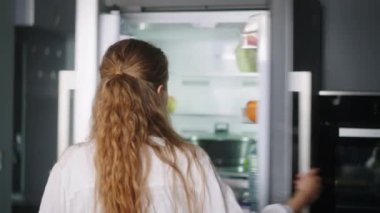 Young woman taking vegetables from fridge and has fun singing and dancing in modern minimalistic kitchen with island. Girl getting tomato from side by side refrigerator and singing to a cucumber.