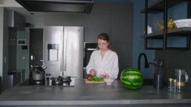Caucasian woman making a salad of vegetables on modern kitchen island. Young female cuts the greens and cooks a salad. Girl cuts cucumbers and tomatoes on chopping board in minimalistic interior.