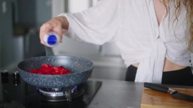 Genç kıvırcık kadın çırpılmış yumurtalarla kızartmak için sıcak tavaya tuz ekliyor. Genç bir kadın gaz ocağında yemek pişiriyor. Kız kahvaltısını modern, minimalist mutfak adasında hazırlıyor..