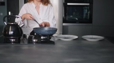 Genç kıvırcık kadın sıcak tavanın tabaklarına çırpılmış domatesli yumurta koyar. Kahvaltıda omlet servisi yapan genç bir bayan. Kız yemeğini modern minimalist mutfak adasında hazırlıyor..