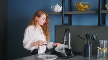 Genç beyaz kadın modern mutfakta lavabonun altında tabak ve bıçak yıkıyor. Ev hanımı mutfak adasında bulaşıkları yıkıyor. Yüksek teknolojili mutfakta bulaşıkları yıkayan kadın.