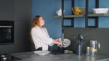Authentic happy caucasian woman washing dishes and dancing in a modern kitchen. Young housewife having fun while doing the washing-up. Girl waving her hair while doing the dishes.