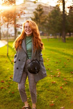 Redhead smiling pretty woman walking in park on sunny autumn day. Elegant female poses in stylish authentic outfit has fun laughing on green lawn with vivid foliage, leaves. Girl enjoys a fall season.