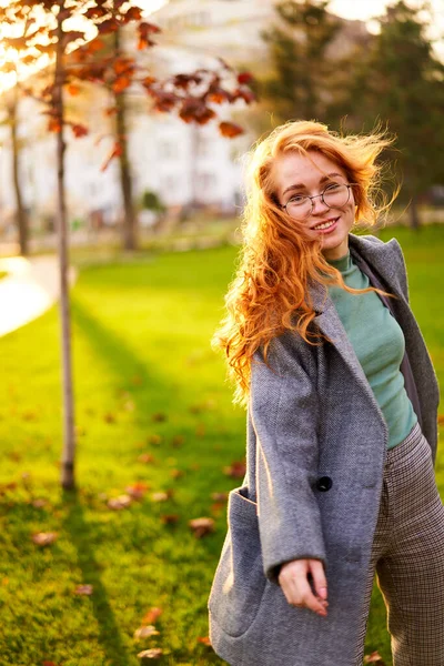 Redhead smiling pretty woman walking in park on sunny autumn day. Elegant female poses in stylish authentic outfit has fun laughing on green lawn with vivid foliage, leaves. Girl enjoys a fall season.