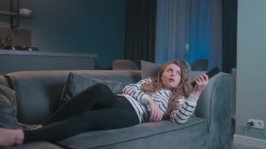 Şirin sarışın kıvırcık kadın televizyonu açıyor, akıllı TV uygulamalarında yol gösteriyor ve evdeki oturma odasındaki kanepede uzanmış kendini eğlendirmek için kanalları seçiyor. Sinematik bir çekim..