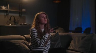 Şirin sarışın kıvırcık kadın televizyonu açıyor, akıllı TV uygulamalarında geziniyor ve evdeki oturma odasında kanepede otururken kendini eğlendirmek için kanalları seçiyor. Sinematik çekim.