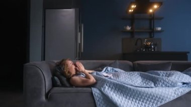 Evdeki kanepede hasta bir kadın öksürüyor ve burnunu battaniyenin altına siliyor. Koronavirüsten muzdarip beyaz bir kadın tecrit edilirken Covid19. Kanepeye uzanmış soğuk kanlı bir kız.