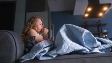 Evdeki kanepede hasta bir kadın öksürüyor ve burnunu battaniyenin altına siliyor. Koronavirüsten muzdarip beyaz bir kadın tecrit edilirken Covid19. Kanepeye uzanmış soğuk kanlı bir kız.