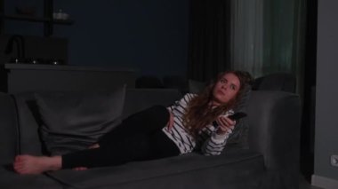 Şirin sarışın kıvırcık kadın televizyonu açıyor, akıllı TV uygulamalarında geziniyor ve evdeki oturma odasında kanepede uzanırken kendini eğlendirmek için kanalları seçiyor. Sinematik çekim.