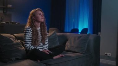 Şirin sarışın kıvırcık kadın televizyonu açıyor, akıllı TV uygulamalarında geziniyor ve evdeki oturma odasında kanepede otururken kendini eğlendirmek için kanalları seçiyor. Sinematik çekim.