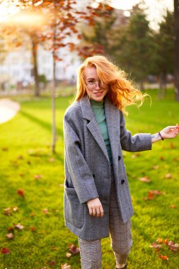 Redhead smiling pretty woman walking in park on sunny autumn day. Elegant female poses in stylish authentic outfit has fun laughing on green lawn with vivid foliage, leaves. Girl enjoys a fall season.