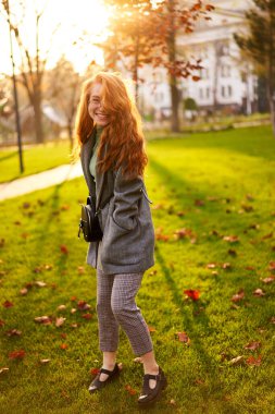 Redhead smiling pretty woman walking in park on sunny autumn day. Elegant female poses in stylish authentic outfit has fun laughing on green lawn with vivid foliage, leaves. Girl enjoys a fall season.
